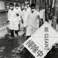 A photograph of five men in white coats wearing masks, holding cleaning supplies on a pavement next to a sign that says 'BE CLEAN'
