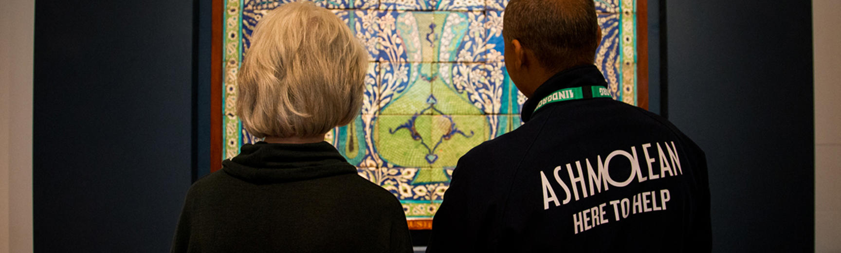 A visitor engagement assistant is telling a visitor some information about the Islamic Tiles in front of them