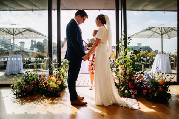 A couple getting married hold hands in front of a sunlit window surrounded by flowers