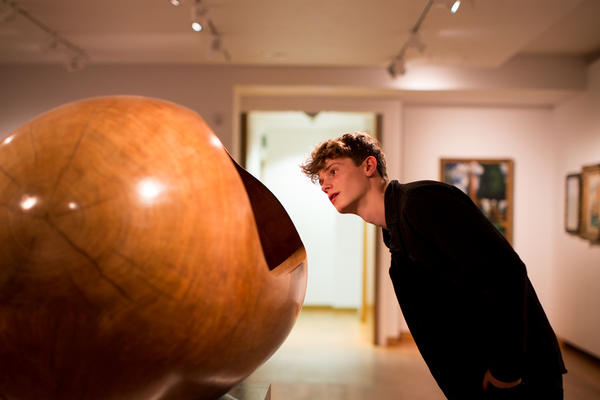 A young man in dark clothes leans towards a smooth wooden carved sculpture by Barbara Hepworth
